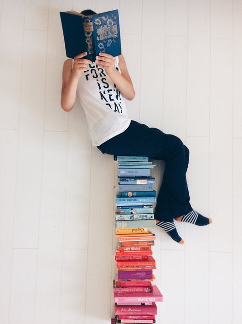 teen sitting on a pile of colorful books, reading 
