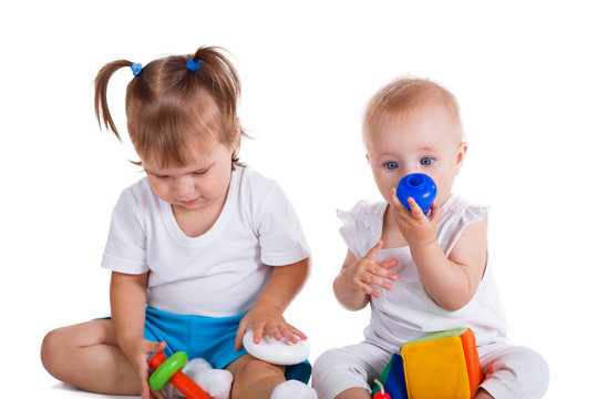 A toddler and a baby sit together playing with blocks.