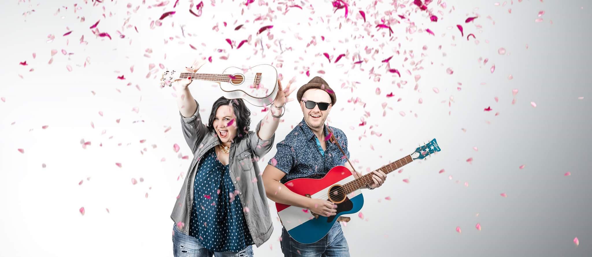 A woman joyfully holds up a ukulele, and a man in hat and sun glasses plays the guitar. Pink confetti explodes behind them. 