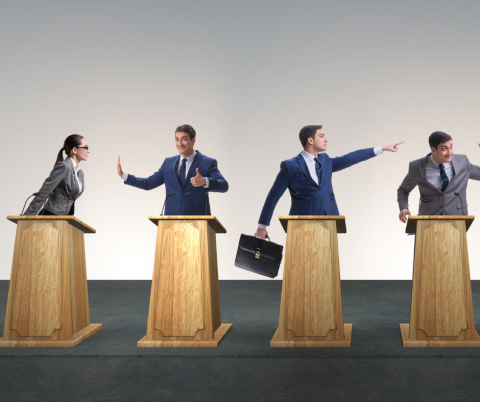 Four individuals at a podium, each person is in a suit pointing angrily at the person next to them.