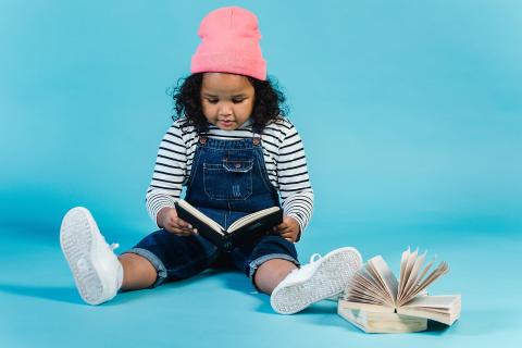 A girl in a pink hat and blue overalls reads a book on her lap. Another open book sits next to her.