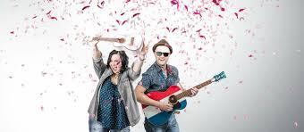 A woman joyfully holds up a ukulele, and a man in hat and sun glasses plays the guitar. Pink confetti explodes behind them. 