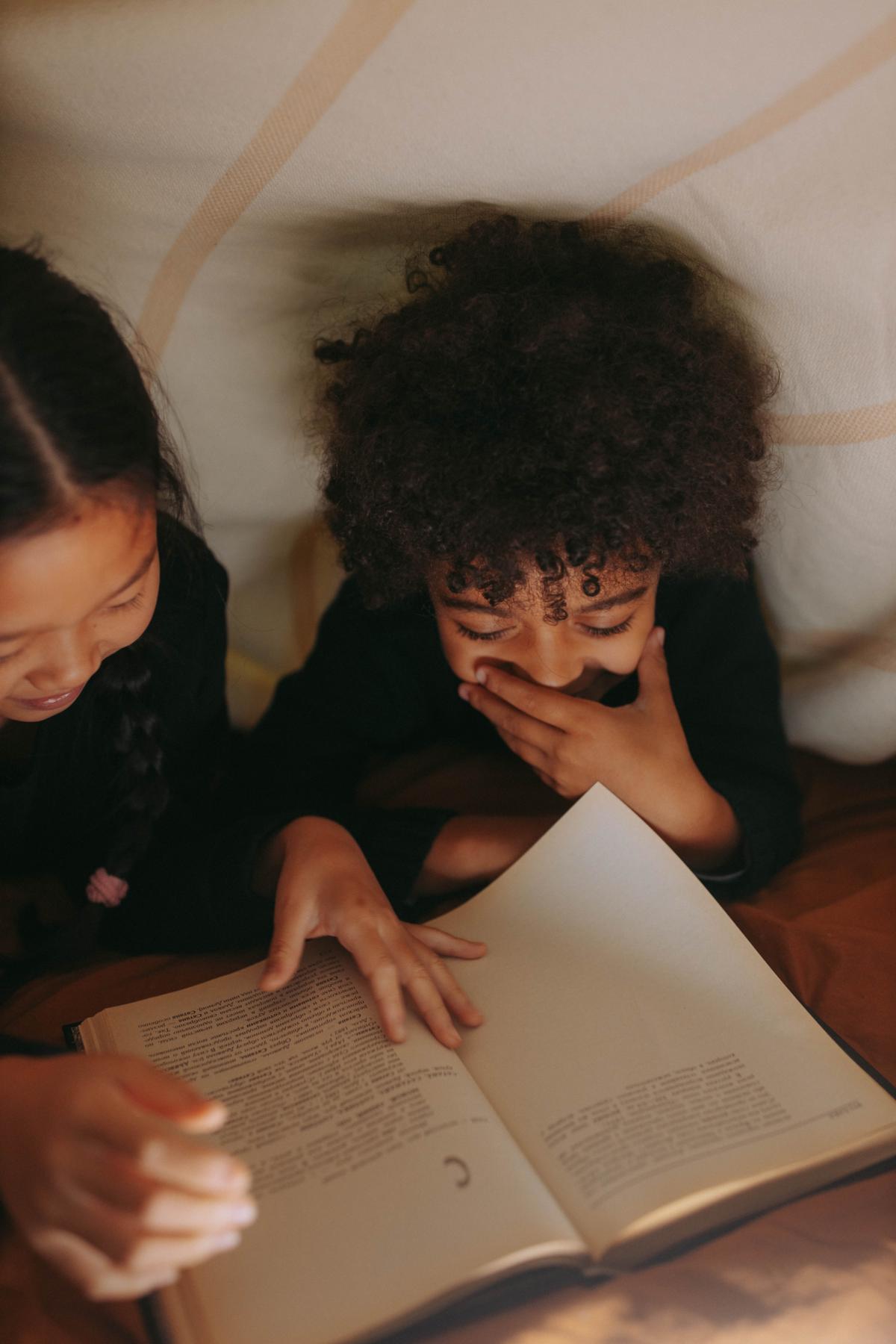 Two children reading in a blanket fort. One child is laughing. 